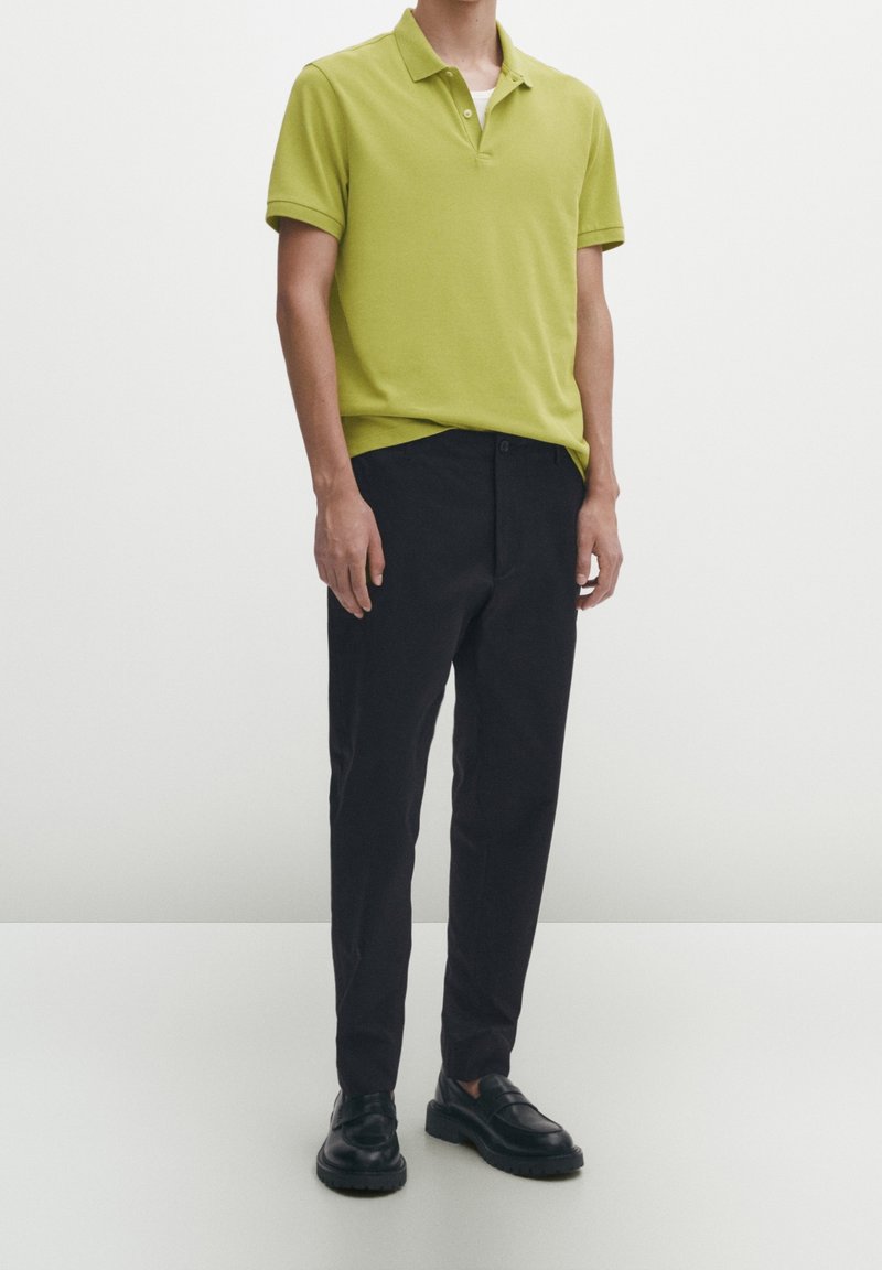 Homme portant un polo manches courtes vert clair, pantalon noir et mocassins noirs, debout devant un fond uni clair.