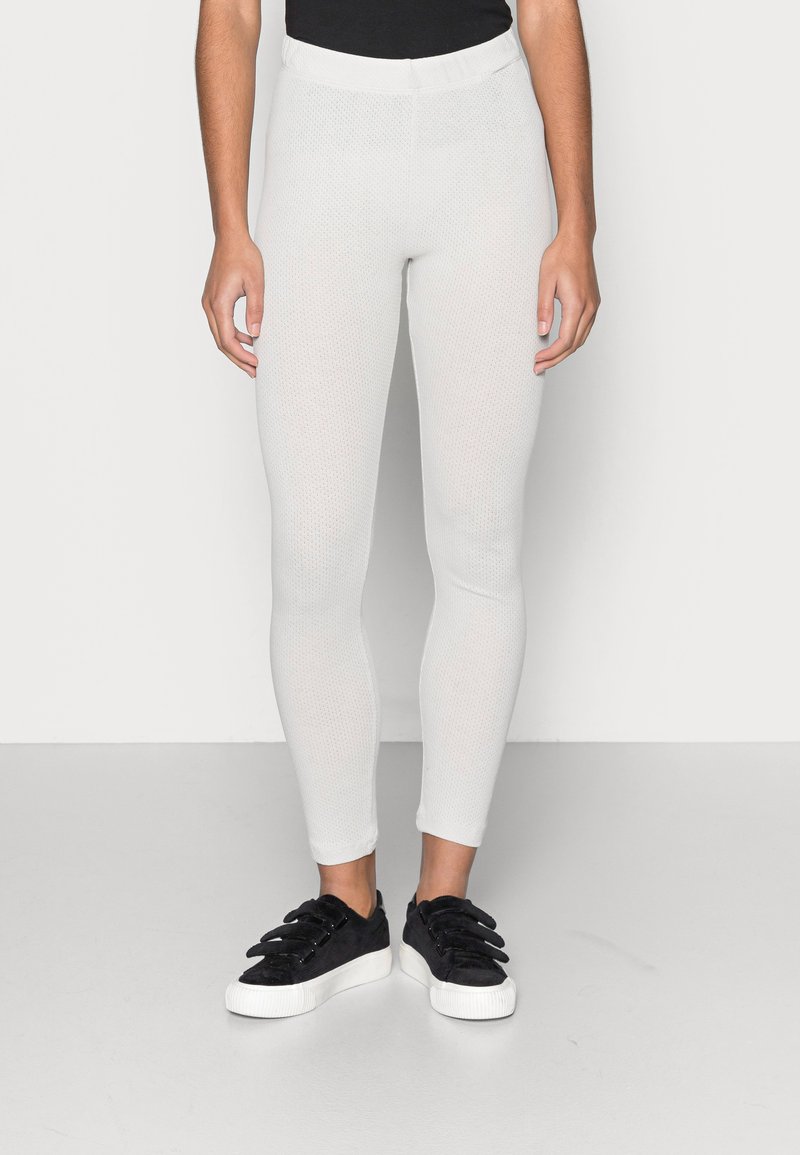 Leggings brancos texturizados com um design ajustado e faixa elástica na cintura, combinados com ténis pretos texturizados com uma sola branca.