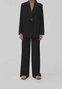 Blazer nero con un unico bottone dorato e due tasche frontali, abbinato a pantaloni neri a gamba larga e scarpe beige. Top a coste con texture sotto.