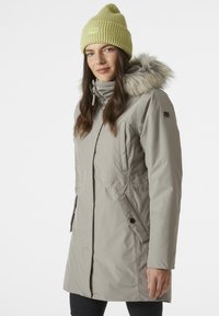 Parka de color gris claro con capucha de piel sintética desmontable, bolsillos con cremallera y cremallera frontal. Combinada con un gorro de punto verde lima.