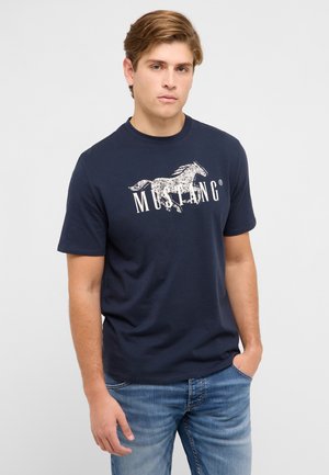 Junger Mann mit hellbraunen Haaren, der ein dunkelblaues Mustang-T-Shirt und hellblaue Jeans trägt, stehend mit einer Hand in der Tasche.