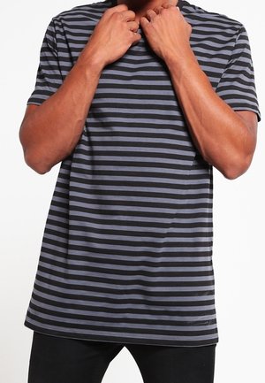 Homme portant un t-shirt à manches courtes rayé horizontalement noir et gris, ajustant le col avec ses deux mains.