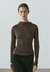 Brown long-sleeve top with a cowl neckline, textured fabric, fitted shape, and gathered detail at the side, paired with black leggings.