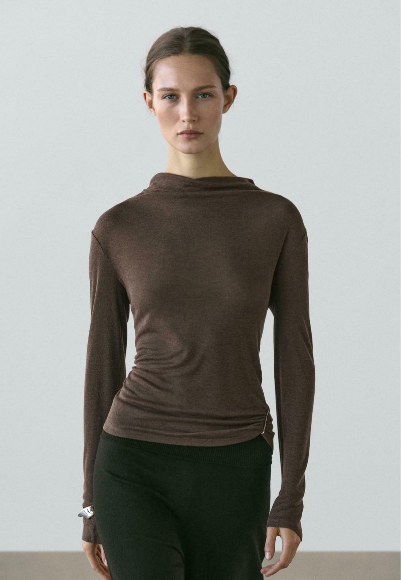 Braunes Langarmshirt mit Wasserfallausschnitt, strukturierter Stoff, figurbetonte Form und gerafftem Detail an der Seite, kombiniert mit schwarzen Leggings.
