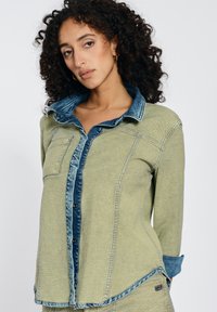 Green shirt with denim accents, featuring a button-up design, collar, and a front pocket. Soft texture and contrasting fabric sections.