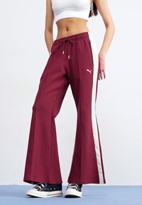En person iført en hvid cropped top og bordeauxfarvede flared sweatpants med en hvid side-stribe står på en lyseblå overflade.