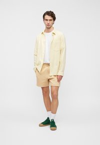 Ανοιχτοκίτρινο πουκάμισο με κουμπιά πάνω από λευκό T-shirt, μπεζ σορτς, πράσινα αθλητικά παπούτσια με καφέ σόλες, φορεμένα με λευκές κάλτσες.