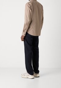 Hombre con camisa de manga larga beige, pantalones negros y zapatillas blancas de pie contra un fondo blanco liso, de espaldas.