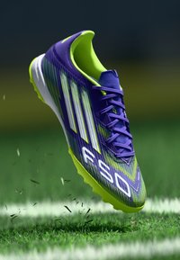 Violets un neona zaļas krāsas futbola čības ar teksturētu virsmu, trim baltām svītrām un izteiksmīgiem saķeres uzgaļiem. Čības šķiet karājas virs zāles.