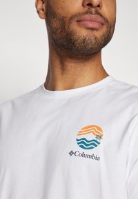 Bílá bavlněná trička s barevným kruhovým logem, které má oranžové vlny a strom, a s názvem značky "Columbia" vytištěným níže.