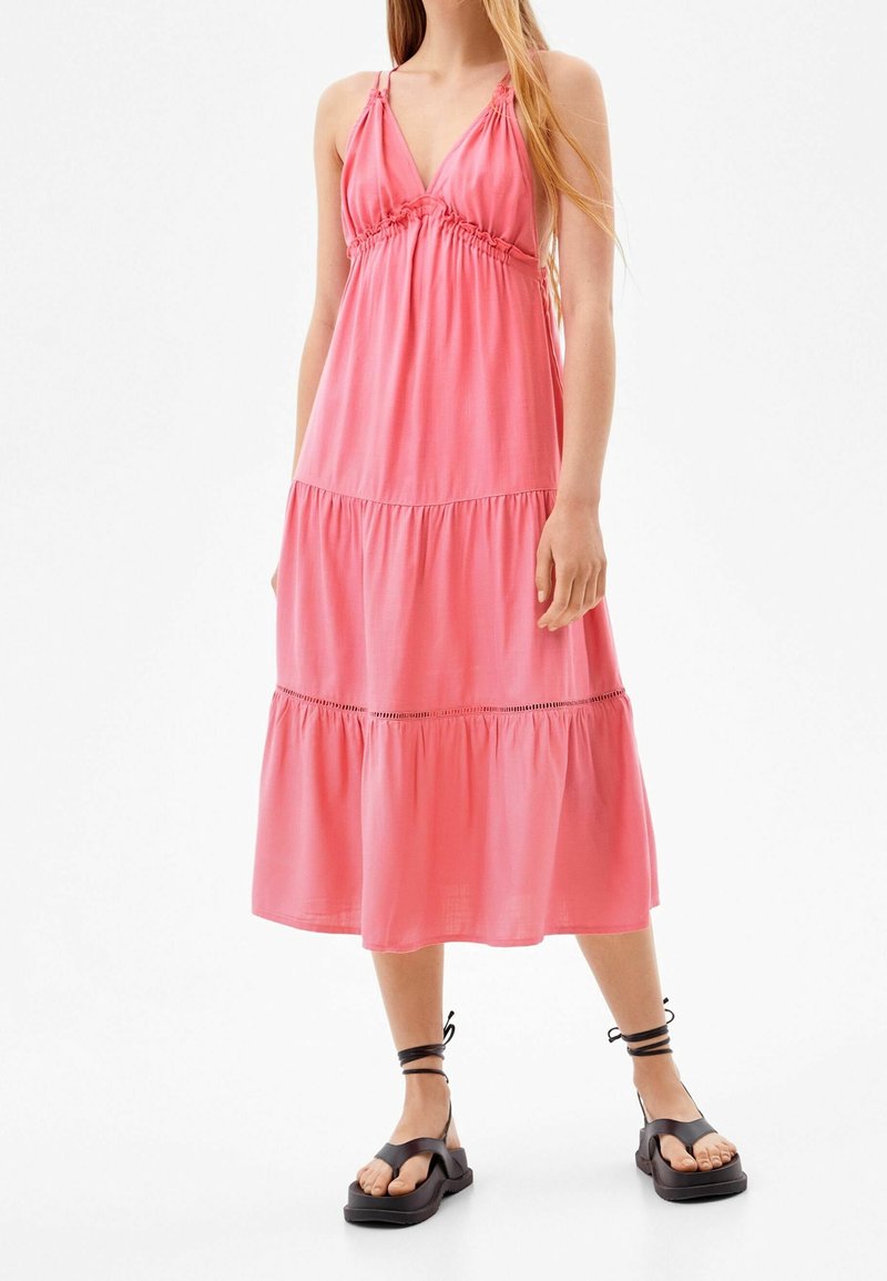 Rosa ärmlös klänning med djup V-ringning, samlad midja, lager-på-lager kjol och en avslappnad passform. Parat med svarta sandaler med ankelband.