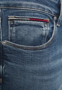 Närbild av en blå denimjeansficka med sömdetaljer och horisontell röd, vit och svart randig trim på den lilla fickan.