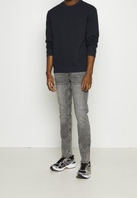 Sudadera de cuello redondo en azul marino, jeans ajustados desgastados en gris, y zapatillas deportivas negras y plateadas. Tejido suave, corte ajustado, costuras visibles y cordones.