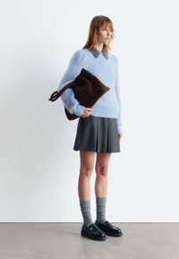 Hellblauer, flauschiger Pullover, grauer Faltenrock, graue knielange Socken, schwarze Loafers und eine braune Tasche mit Schulterriemen. Minimalistischer Hintergrund.