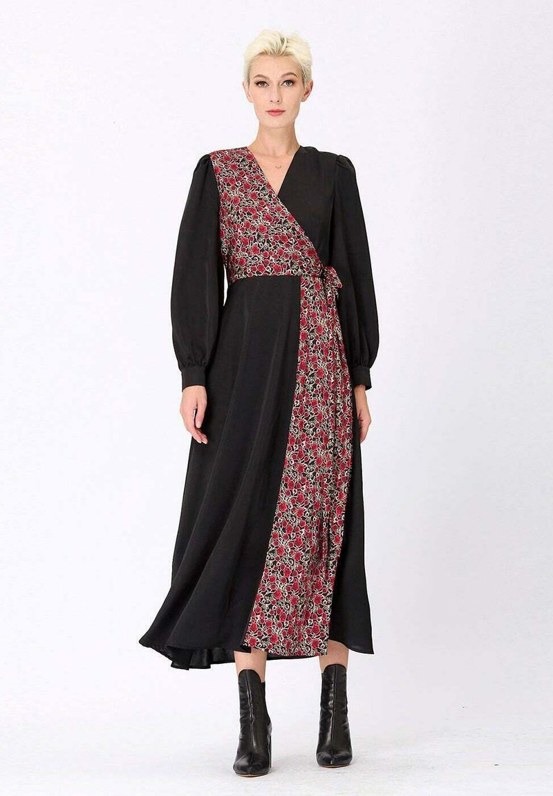 Robe noire enveloppante avec un motif floral sur le côté gauche. Présente des manches longues bouffantes, un décolleté en V, et une longueur jusqu'aux chevilles. Portée avec des bottes noires.