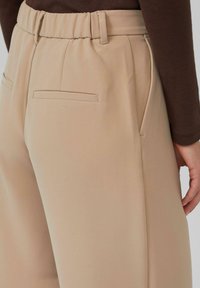 Beige maßgeschneiderte Hosen mit glatter Textur, elastischem Bund und zwei Gesäßtaschen, die ein sauberes und minimalistisches Design präsentieren.