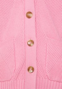 Närbild av en rosa stickad kardigan med tre stora bruna knappar och ett strukturerat chevron-stickningsmönster på fickorna.