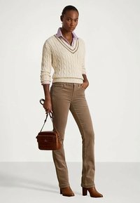 Kobieta w kremowym swetrze w sploty noszącym na lawendowej koszuli, brązowych spodniach z kordurowej tkaniny, brązowych butach, trzymająca małą brązową torebkę.