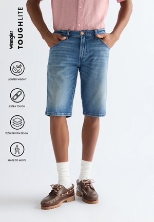 Man die lichtblauwe denim shorts, een roze short-sleeve shirt, witte sokken en bruine leren schoenen draagt, staand met handen in de zakken.