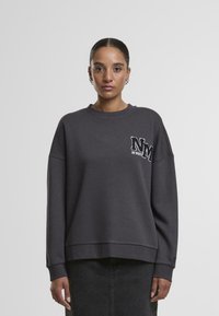 Sweatshirt gris foncé avec un col rond, des épaules tombantes et un logo "NM" brodé texturé sur la poitrine. Tissu doux, coupe décontractée.