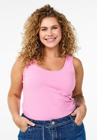 Rosa linne med rundad halsringning, tillverkat av mjukt tyg, kombinerat med blå högmidjade denimjeans. Modellen står med händerna i fickorna.