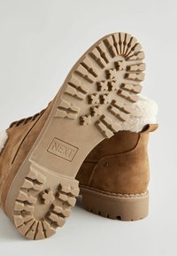 Bota de ante marrón claro con un forro blanco esponjoso. Suela de goma texturizada pesada con un dibujo en el dibujo. Logo "NEXT" grabado en el lateral.