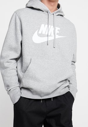 Person trägt einen hellgrauen Nike-Kapuzenpullover mit weißem Logo, Hände in den Taschen, kombiniert mit schwarzen Hosen vor einfarbigem Hintergrund.