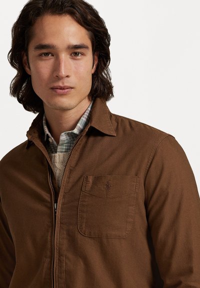 Chaqueta marrón con cremallera y cuello, con un bolsillo en el pecho izquierdo. La tela parece suave con costuras visibles. Se lleva sobre una camisa de cuadro.