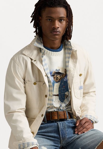 Beige vászondzseki nagy zsebekkel, egy kék és fehér kockás gombos ing és egy bears mintával ellátott texturált kötött pulóver fölött.