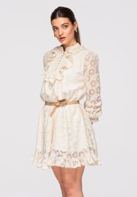 Femme portant une robe en dentelle crème avec un devant à volants, des manches longues transparentes, une taille cintrée par une ceinture beige, debout devant un fond gris clair.