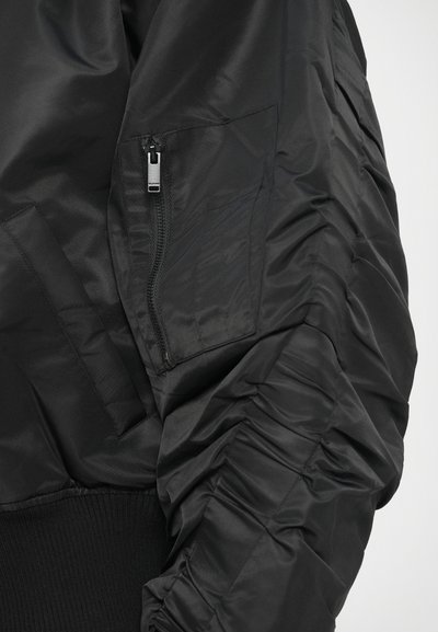 Juodas bombonešio stiliaus švarkas, pagamintas iš lygaus, blizgaus audinio, su gofruotomis rankogaliais, tekstūruota surenkta siūle rankovėse ir sagomis užsegama kišene ant rankovės.