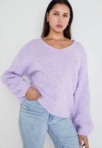Lavendelkleurige gebreide trui met een V-hals, losse pasvorm en wijde mouwen, gecombineerd met lichtblauwe jeans. Zachte textuur, geribbelde afwerking aan de randen.