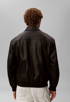 Homme aux cheveux bouclés portant une veste en cuir marron foncé et un pantalon blanc, vu de dos sur un fond gris uni.