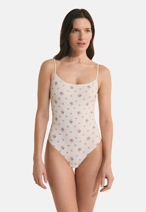Femme portant un maillot de bain une pièce côtelé de couleur claire avec un motif floral et de fines bretelles, debout devant un fond blanc uni.