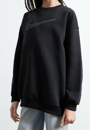 Osoba w czarnej bluzie Nike z kropkowanym logo Swoosh, w jasnoniebieskich dżinsach, na jednolitym jasnym tle.