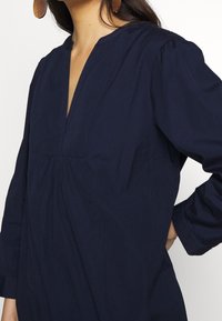 Marinblå bomullstunika med bred halsringning, lång ärm och rynkdetaljer vid bysten. Slät textur med en avslappnad passform.