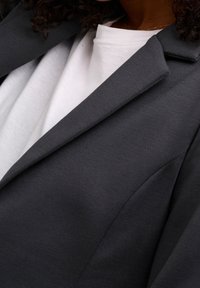 Blazer gris hecho de tela texturizada, con solapas, hombros estructurados y una silueta ajustada sobre una camisa blanca.