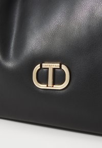 Zwarte leren tas met textuur en opvallende goudkleurige hardware met de merknaam "TWINSET." Gladde afwerking en ronde vorm.