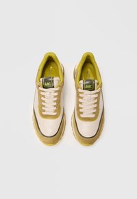 Sneakers met een beige leren en olijfgroene suède bovenkant, voorzien van een zwarte accentlijn, witte veters en een gele rubberen zool.