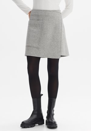 Femme portant une jupe portefeuille grise avec une grande poche, des collants noirs, des bottes noires épaisses et un haut blanc à manches longues, debout les mains sur les hanches.