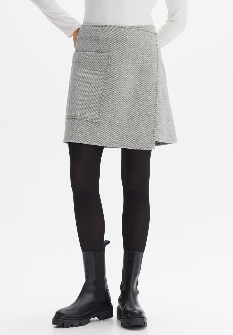 Femme portant une jupe portefeuille grise avec une grande poche, des collants noirs, des bottes noires épaisses et un haut blanc à manches longues, debout les mains sur les hanches.