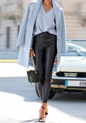 Kobieta spacerująca na zewnątrz, ubrana w jasnoniebieski płaszcz, niebieski sweter z dekoltem w serek, czarne skórzane spodnie, czarne szpilki, złote łańcuszki i trzymająca czarną torebkę.