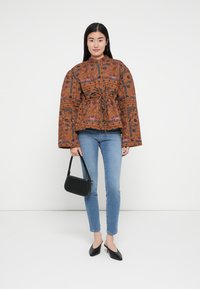 Bruine jas met patroon en een getailleerde taille, brede mouwen en bloemmotieven. Draagt over een blauwe skinny jeans en gecombineerd met een zwarte handtas.