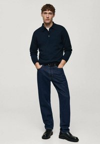 Maglietta polo in maglia navy con chiusura a due bottoni, abbinata a jeans in denim scuro e scarpe nere stringate. Design semplice, vestibilità casual.