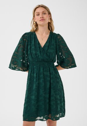 Femme portant une robe texturée vert foncé avec des manches bouffantes trois-quarts transparentes et un décolleté en V, debout sur un fond clair uni.