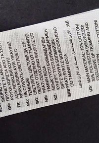 Etykieta pielęgnacyjna ubrania z instrukcjami prania i składem materiału w kilku językach na białej tkaninie na ciemnym tle.