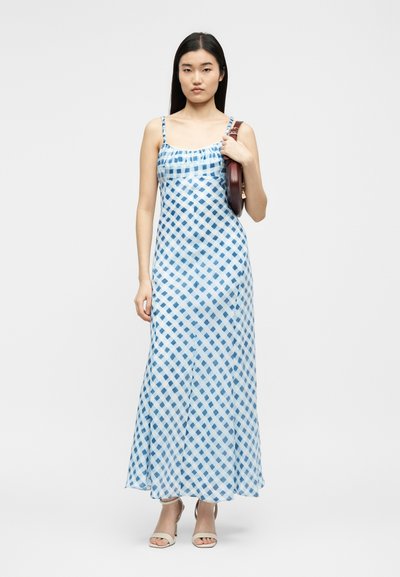 Γυναίκα στέκεται φορώντας μπλε-λευκό καρό αμάνικο μακρύ φόρεμα, μπεζ σανδάλια με τακούνι και κρατώντας καφέ τσάντα στον ώμο της.