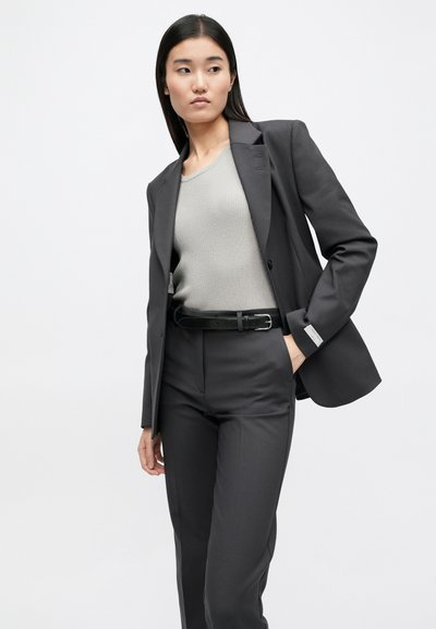 Σκούρο γκρι κοστούμι με προσαρμοσμένο σακάκι και ταιριαστά παντελόνια, συνδυασμένο με ένα ανοιχτό γκρι ριγέ τοπ και μια μαύρη ζώνη.