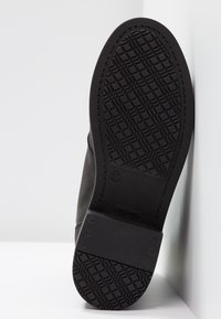 Semelle de chaussure en caoutchouc noir présentant un motif texturé avec des carrés et des lignes, conçue pour offrir une adhérence, montrée en angle contre un fond clair.