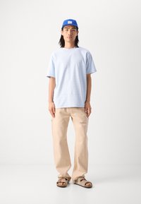 Lyseblå kortærmet skjorte med diskrete striber, beige brede bukser og brune sandaler. En blå kasket med en hvid label fuldender looket.